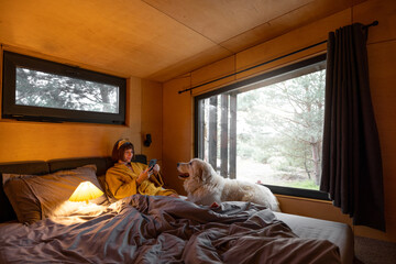 Wall Mural - Woman uses phone while lying in bed with her cute dog in tiny bedroom of wooden cabin on nature