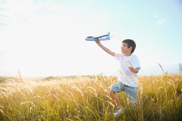 Wall Mural - Cute happy cheerful child running fastly along grassy hill at countryside holding big toy plane in hand. Boy playing during sunset time in evening. Horizontal color photography.