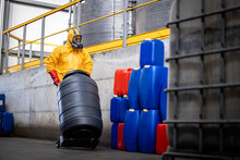 Worker Wearing Protective Suit And Gas Mask While Moving Dangerous Chemicals At Modern Chemical Plant. In Background Large Tanks Reservoirs With Acid.