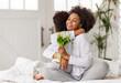 © JenkoAtaman - Happy mother's day! Afro american family happy child daughter congratulates mom on holiday, hugs her and gives bouquet of flowers