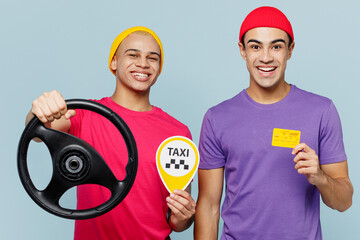 Young fun smiling happy couple two friends men wear casual clothes together hold taxi card sign steering wheel credit bank card driving car isolated on pastel plain light blue cyan background studio.