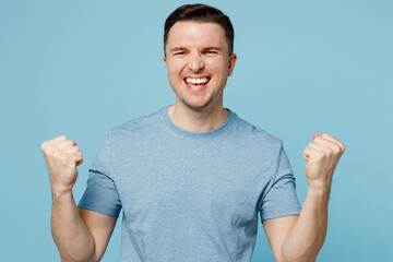 Wall Mural - Young overjoyed happy caucasian man wear casual t-shirt doing winner gesture celebrate clenching fists say yes isolated on plain pastel light blue cyan background studio portrait. Lifestyle concept.