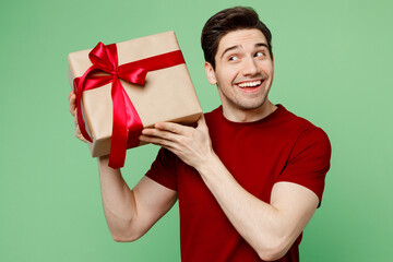 Poster - Young smiling happy fun cheerful man he wearing red t-shirt casual clothes holding present box with gift ribbon bow isolated on plain pastel light green background studio portrait. Lifestyle concept.