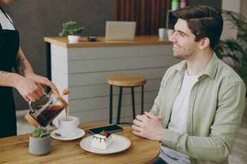 Wall Mural - Young smiling happy man wear shirt casual clothes talk with waiter pouring tea from teapot into cup sitting alone at table in coffee shop cafe restaurant indoors rest relax during free time inside.