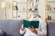 Animal reader with a book. Man with a funny rabbit face reading in living room at home. Bizarre guy wearing white shirt, bunny mask and eyeglasses sitting on sofa with interesting novel or fairy tale