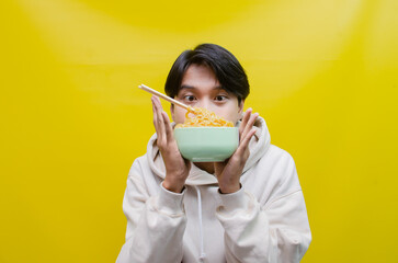 Closeup an Asian man in a beige hoodie eats instant noodles using chopsticks and a bowl, isolated over a yellow background. 