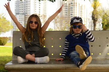 Cool kids. A boy and a girl are sitting on a bench, children in sunglasses hid in the shade. Brother and sister, best friends.