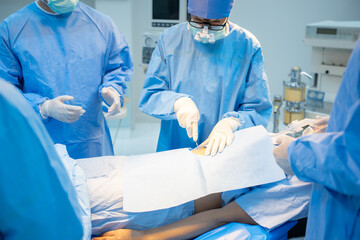 Front view of a Caucasian female surgeon with team, in blue surgical uniform, face mask, and medical loupes, standing surgery on the patient's stomach through a surgical drape in the operating room.