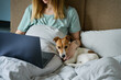 Woman with cute dog relaxing in bed at morning and use laptop. Comfortable work from home for freelancer. Spending time together, pet affection
