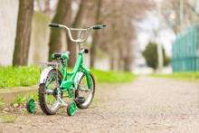 A Green Bicycle For Children With Additional Wheels At Road In Park