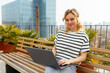 © BONNINSTUDIO/Stocksy - Blonde woman using laptop on bench on building terrace