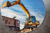Fototapeta  - A wheeled excavator loads a dump truck with soil and sand. An excavator with a high-raised bucket against a cloudy sky View from the trench. Removal of soil from a construction site or quarry.