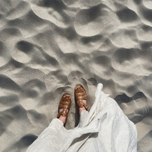 Women Feet On Desert Beach Sand. Aesthetic Summer Vacation Background. Woman In White Dress And Leather Sandals On The Dune Sand. Top View