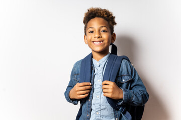 Portrait of a cute 5-6 year old boy with dark skin and afro hair posing on a white background. The boy is carrying a backpack. Infant school concept, children begin the primary stage.