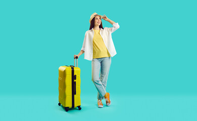 Full body happy young woman in summer hat, casual shirt and blue jeans standing with yellow suitcase isolated on turquoise background, looking away and smiling. Holiday, vacation, traveling concept