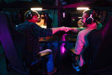 Two male gamers giving fist bump while playing video game together in cybersport club