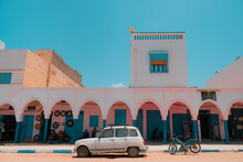 Mirleft, Morocco - Colorful Market Exterior With Blue Doors And Windows, Pink Walls, White Arches. Vintage Vehicles Parked Outside: A White Renault 4 And An Old Blue Motorcycle. Spanish Architecture.
