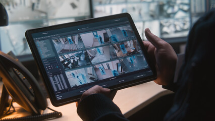 Male security officer controls CCTV cameras in office, uses digital tablet and computers with surveillance camera footage playback on screens. High tech security system. Monitoring and social safety.