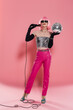 Full length of trendy drag queen in sunglasses and gloves holding microphone and disco ball on pink background.