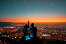 Rear View Silhouettes Of A Couple Sitting On The Top Of The Hill Looking And Pointing Out At Shooting Star Over The City In The Sky