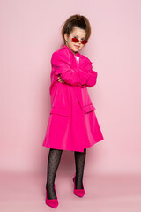 Wall Mural - Portrait of a little girl in an oversized pink blazer and a slim stylish red glasses. Child wearing adult clothes. Isolated on pink background.