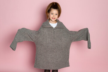 Portrait of a little girl in an oversized brown sweater with a hanging sleeves. Child wearing adult clothes. Isolated on pink background.