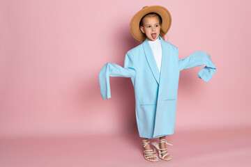 Wall Mural - Portrait of a little girl in an oversized adult blue blazer and a round brown hat. Child wearing adults clothes. Isolated on pink background.