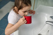 Close-up portrait of 5-6 years old Caucasian little child girl in white pajamas, rinsing her mouth with water while brushing teeth, standing by ceramic sink in the home bathroom. Oral care and hygiene