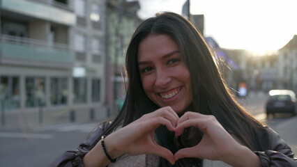 One happy young woman doing heart symbol with hands standing in city street during sunset time. Portrait face of a person makes heart shape gesture