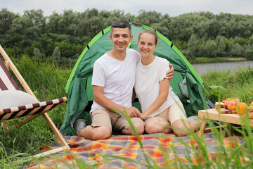 Wall Mural - Smiling happy optimistic woman and man wearing casual clothing sitting at tent near the river looking at camera having picnic enjoying spring days.