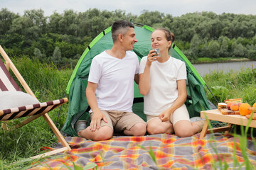 Wall Mural - Tasty coffee on nature. Joyful smiling woman and man wearing casual clothing sitting at tent near the river, drinking coffee, enjoying aromat, resting, relaxing.