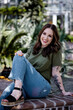 Fashionable brunette Caucasian female with tattoos wearing a green shirt and blue jeans around green plants