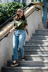 Fashionable brunette Caucasian female with tattoos wearing a green shirt and blue jeans around green plants