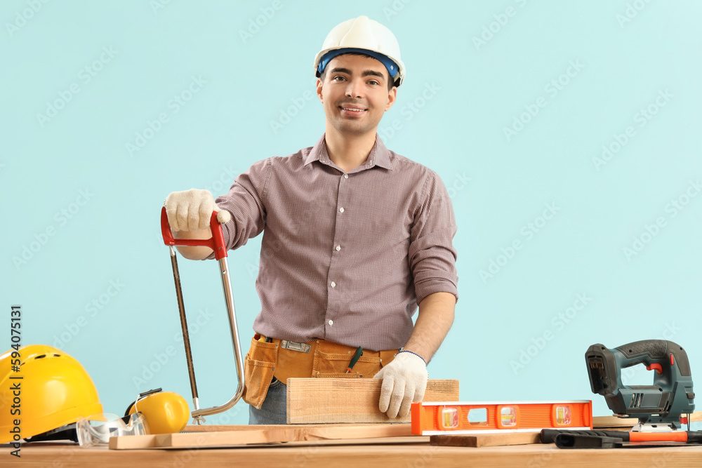 Young carpenter with hacksaw and wooden plank at table on blue background