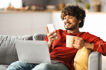 Wall Mural - Handsome Indian Guy Relaxing At Home With Smartphone, Laptop And Coffee