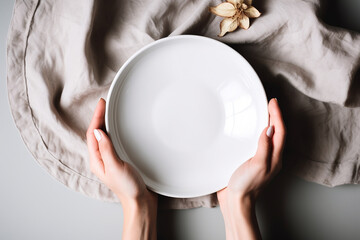 Wall Mural - Woman's hands holding white empty plate on the light table with linen napkin decorated with dry flower. Generative AI