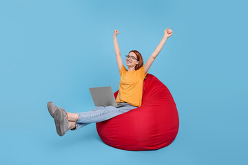 Poster - Happy young woman with laptop sitting on beanbag chair against light blue background, space for text