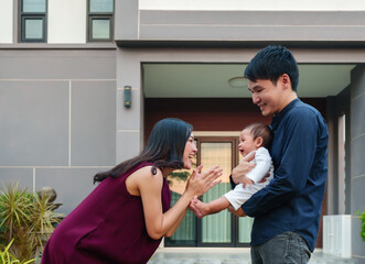 Wall Mural - happy family, cheerful father and mother holding and playing with newborn baby while standing in front of their house