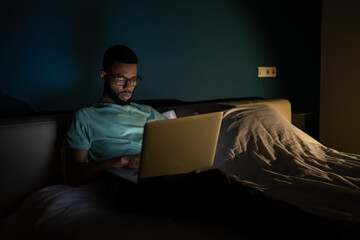 Focused African American man wearing glasses looking at laptop screen working on project until night, lying in bed with laptop. Black guy freelance IT developer coding at night to meet deadline