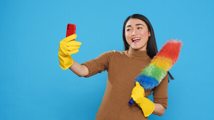 Wall Mural - Smiling housekeeper holding colorful feather dust while taking selfie with mobile phone, standing in studio over blue background. Cheerful maid using high-quality detergents and rubber gloves