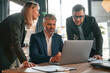 © standret - Senior man is sitting by laptop, employees are helping. Group of business people are working in the modern office