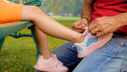 Father ties his daughter's shoes - childcare and parenting. Closeup shot of a caring dad helping his little girl to tie her shoelaces while she is sitting on a bench in the park