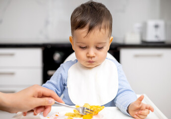 adorable baby boy kid eating peach pancake,mother hand feeding child. wide open mouth hungry cute boy with white bib modern kitchen home house interior.isolated infant food diversification high