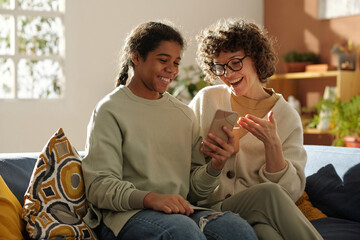 Wall Mural - Happy mom spending time with her adopted daughter at home, they sitting on sofa and watching photos on mobile phone