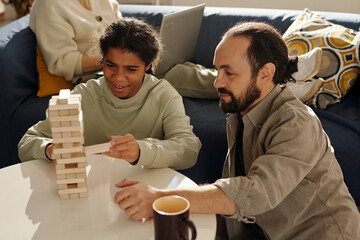 Wall Mural - Teenage girl playing jenga with her foster dad in the living room with mother working on laptop in background