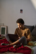 © pressmaster - Vertical image of teenage boy using laptop while sitting on his bed and eating chips