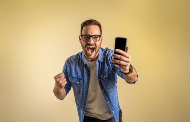 Wall Mural - Portrait of cheerful male entrepreneur with smart phone screaming ecstatically and pumping fists. Young man reading message and celebrating success over beige background