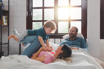 Dad and mom playing and bonding with cheerful kid in the morning. Caucasian mother lifting his cute little son in the air to pretend to fly like a plane or superhero with arms out on a bed at home.