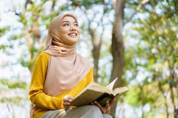 Wall Mural - Happy cheerful Asian beautiful Muslim woman reading a book - Koran under the tree. Female muslim student studying an Islam Koran.