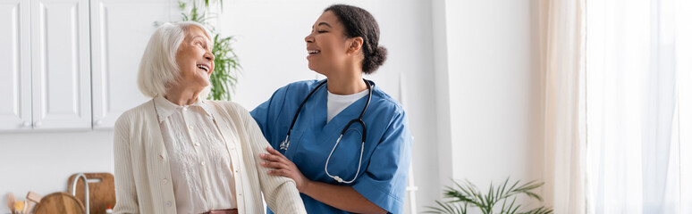 happy multiracial nurse in uniform laughing with retired woman at home, banner.